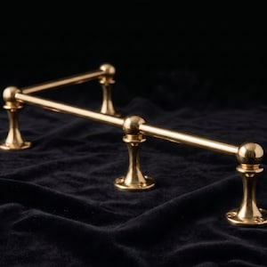 May include: A polished brass gallery rail featuring a corner design with three vertical supports. The horizontal rods are connected by spherical joints, and each base has a circular mounting plate with visible screw holes for installation.