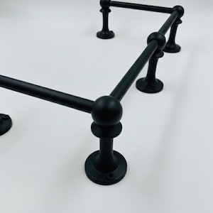 Könnte beinhalten: Schwarzer Metall-Handtuchhalter mit geometrischem Design. Der Halter hat abgerundete Verbindungen und eine matte Oberfläche. Die Basis ist kreisförmig und das Design eignet sich für ein Badezimmer oder eine Küche. Maße in Zentimetern.