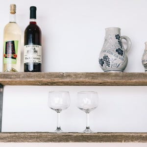 Peut inclure: Une étagère en bois rustique avec des supports métalliques présente des bouteilles de vin, des pichets décoratifs et des verres à vin. L'étagère est en bois de récupération et est fixée à un mur blanc. Les bouteilles ont des étiquettes et les pichets des motifs.