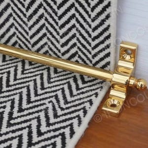 Puede incluir: Un retenedor de ventana de latón pulido con un extremo de bola decorativo y placas de montaje. El retenedor está colocado sobre una superficie de madera junto a una alfombra con estampado de chevrón en blanco y negro.