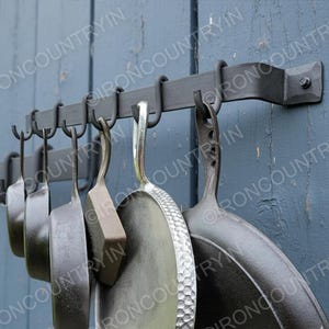 May include: A black wrought iron pot rack with several cast iron frying pans suspended from it. The rack is mounted on a weathered blue wooden surface. The pans vary in size and shape, creating a rustic kitchen aesthetic.