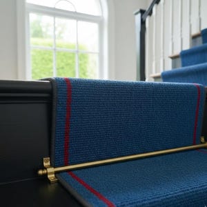 May include: A blue stair runner with a textured weave and two vertical red stripes running along the length. The runner is secured to the black wooden stairs with a horizontal brass stair rod featuring decorative finials at each end.