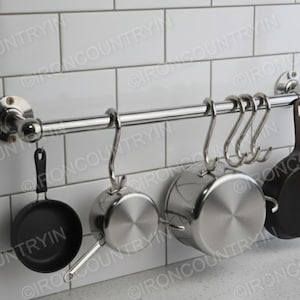 Peut inclure: Un présentoir à casseroles en acier inoxydable avec six crochets, contenant diverses casseroles et poêles. Le présentoir est monté sur un mur carrelé blanc. Les poêles comprennent des casseroles en acier inoxydable et des poêles en fonte noire.