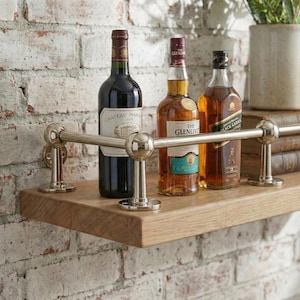Puede incluir: Una estantería de madera con tres botellas de alcohol. Se exhiben una botella de vino tinto, una botella de whisky escocés y una botella de whisky. La estantería está sostenida por una barra de metal con detalles de bolas decorativas.