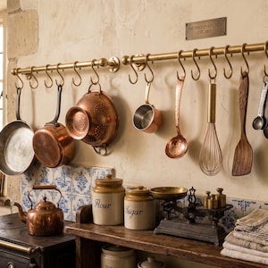 May include: A brass kitchen rail with S-hooks holding copper pans, a colander, a small saucepan, a ladle, a whisk, a wooden spatula, measuring spoons, and a corkscrew. Below, a copper kettle, ceramic flour and sugar jars, and a scale.