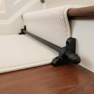 Puede incluir: Una barra negra para alfombra de escalera instalada en una escalera. La barra es de metal y tiene terminales decorativos. La alfombra de la escalera es de color crema y está asegurada por la barra. Las escaleras tienen pasamanos y contrahuellas de madera. La barra mide unos 76 cm.