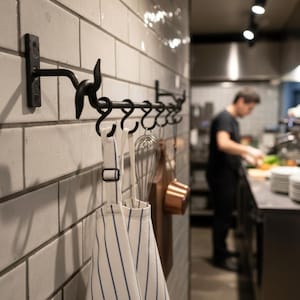 May include: A black metal kitchen rack with hooks is mounted on a white tiled wall. Hanging from the hooks are a striped apron, a whisk, a wooden cutting board, and a copper pot. A person is working in the background.