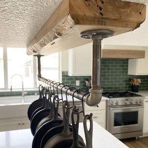 Puede incluir: Un estante para ollas de estilo industrial rústico suspendido del techo. El estante presenta una gruesa repisa de madera con borde natural sostenida por tuberías metálicas. Una fila de sartenes de hierro fundido cuelga, mostrando una solución de almacenamiento de cocina funcional.