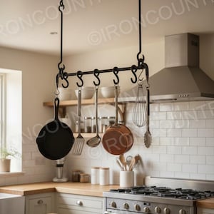 Op de afbeelding: Zwart smeedijzeren pottenrek dat aan het plafond hangt en diverse keukengerei en kookgerei bevat. Inclusief een gietijzeren koekenpan, koperen pan en roestvrijstalen gereedschap. Een functionele en decoratieve opbergoplossing voor de keuken.