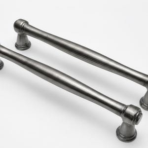 Może przedstawiać: Dwa uchwyty do szafek w stylu antycznym z ciemnoszarym wykończeniem. Każdy uchwyt ma cylindryczny środek i dekoracyjne końcówki. Uchwyty są przeznaczone do łatwego montażu i dodają uroku vintage każdej szafce.