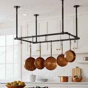 Op de afbeelding: Zwarte metalen pottenrek aan het plafond met zes koperen pannen. De keuken heeft witte kasten, een raam en een houten snijplank. De pannen zijn in verschillende maten en vormen.