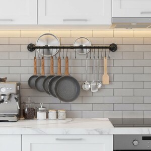 May include: A kitchen scene featuring a black metal pot rack with hanging cookware. Gray granite-style frying pans with wooden handles, stainless steel utensils, and lids are suspended. The backdrop is a white tiled wall and white cabinets.