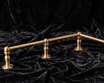 May include: A brass-toned metal rail featuring a smooth cylindrical rod supported by three vertical posts with rounded finials and circular mounting bases, designed for wall installation with visible screw holes on each base plate.