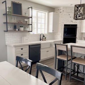 Könnte beinhalten: Eine moderne Küche mit weißen Schränken, einem schwarzen Geschirrspüler und einem Bauernhaus-Waschbecken. Die Küche verfügt über eine weiße Arbeitsplatte, einen schwarzen Wasserhahn und eine Holzinsel mit Barhockern. Ein Holzregal mit Dekoration ist an der Wand befestigt.