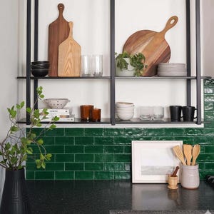 May include: A kitchen shelf displays wooden cutting boards, bowls, glasses, and mugs. A small potted plant and various kitchen utensils are also visible. The shelf is mounted against a green tiled backsplash, with a black countertop below.
