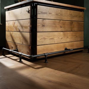 Puede incluir: Una barra de madera rústica con un acabado natural, con un reposapiés y un marco de tubería de metal negro. La barra tiene un diseño multicapa con vetas de madera visibles y está sobre un suelo de madera.