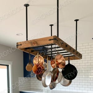 May include: A wooden pot rack suspended from the ceiling with black metal supports. Various copper and stainless steel pots and pans hang from the rack, showcasing a kitchen storage solution. The rack is mounted above a blue cabinet.