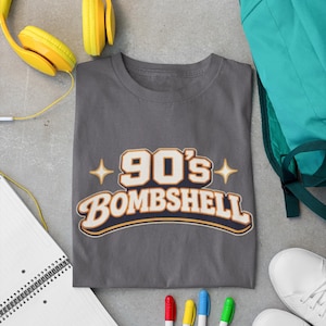 May include: A gray t-shirt with the text "90's Bombshell" in a retro design. The image also includes yellow headphones, a teal backpack, white sneakers, a notepad, and colorful markers.