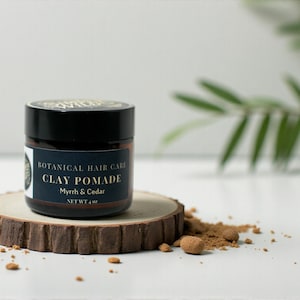 May include: A black jar of clay pomade with a dark blue label that reads "Botanical Hair Care Clay Pomade Myrrh & Cedar NET WT 113 g." The jar sits on a small wooden slice with brown granules and small round objects scattered around it.