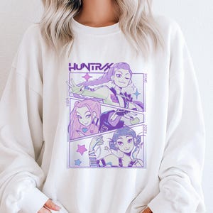 May include: White sweatshirt featuring a comic-style graphic with three anime-style characters and the word "HUNTRA" in purple. The graphic includes stars and the names "MIA," "RUMI," and "ZOEY." The sweatshirt has long sleeves and a crew neck.