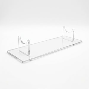 Puede incluir: Un soporte de exhibición de acrílico transparente con una base rectangular y dos soportes verticales, cada uno con un recorte semicircular curvo en la parte superior diseñado para sostener objetos alargados horizontalmente.