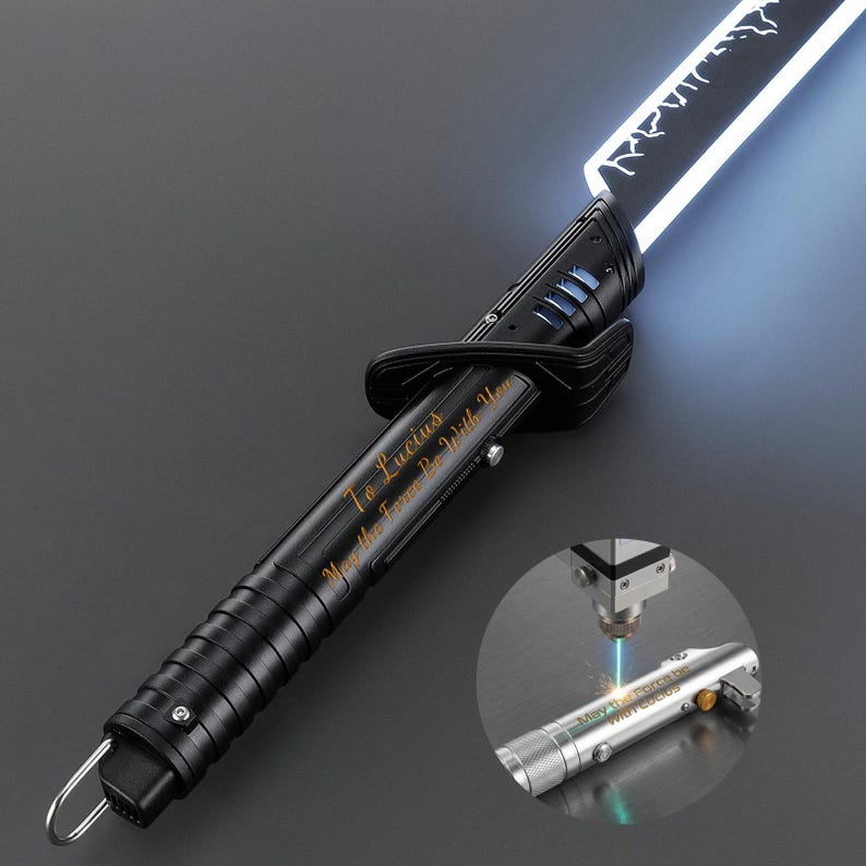Op de afbeelding: Zwart lichtzwaard met een helder wit blad. De handgreep is gegraveerd met de tekst "To Lucius, May the Force Be With You." Een kleinere afbeelding toont het graveerproces op een zilveren lichtzwaard.