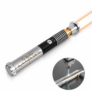 Puede incluir: Un sable de luz plateado y negro con hoja naranja, con una empu&ntilde;adura met&aacute;lica grabada con el texto "To Lucius May the Force Be With You". Una imagen circular insertada muestra una herramienta de grabado l&aacute;ser marcando el texto.