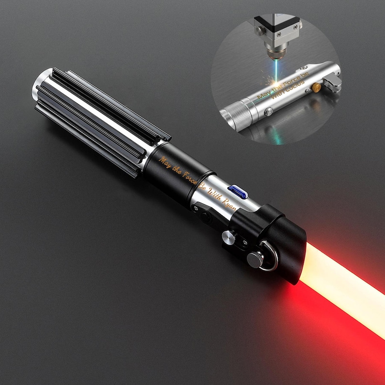 May include: A black and silver lightsaber with a red blade. The hilt has a ribbed design and the text "May the Force Be With Ryan" is engraved on the side. A smaller, similar lightsaber is being laser engraved with the same text.