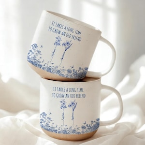 Puede incluir: Dos tazas de cerámica blanca apiladas. Cada taza tiene un diseño floral azul y el texto "IT TAKES A LONG TIME TO GROW AN OLD FRIEND". Los nombres "Sophia" y "Andrea" también están impresos. Las tazas tienen una base rústica sin esmaltar.