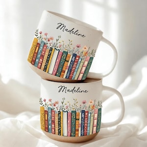 Könnte beinhalten: Zwei weiße Keramikbecher, die übereinander gestapelt sind, jeder mit einem einzigartigen Design. Die Becher zeigen eine Reihe bunter Bücher mit floralen Akzenten und dem Namen "Madeline" in Schreibschrift. Die Becher haben einen beigen Boden und einen weißen Henkel.