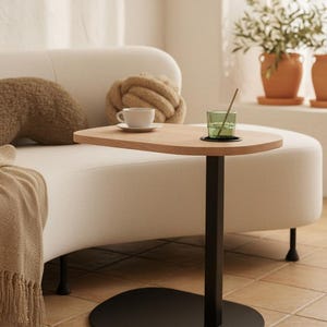 May include: A modern side table with a light wood top and a black metal base. The table has a unique shape and is designed to be placed next to a sofa. A cup and a glass are on the table.
