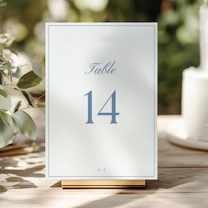 May include: A rectangular white table number card featuring a thin blue border, the word Table in blue script, and the number 14 in a large blue serif font. The card is held upright by a gold metal stand and includes the small letters S T.