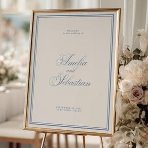 Op de afbeelding: Een rechthoekig welkomstbord voor een bruiloft in een dunne gouden lijst met een crèmekleurige achtergrond en een blauwe dubbele rand. De tekst luidt: WELCOME to the wedding of Amelia and Sebastian SEPTEMBER 19, 2027 LAKE COMO | ITALY in blauw.