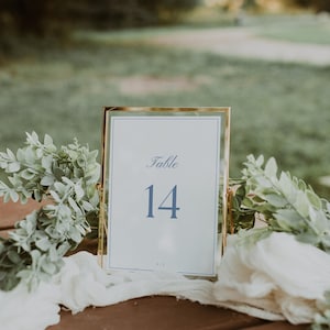 Puede incluir: Una tarjeta con el número de mesa con marco dorado que muestra la palabra "Table" y el número "14" en azul. La tarjeta está sobre una superficie de madera, con vegetación y tela blanca en el fondo.