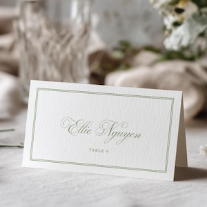 May include: A white folded place card with a textured surface featuring a thin sage green double-line border. The name Ellie Nguyen is printed in a dark green elegant script font, with the text TABLE 9 centered in a simple serif font below.