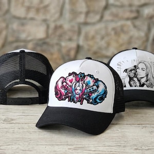 Könnte beinhalten: Drei Trucker-Caps mit schwarzem Netzrücken und schwarzem Schirm. Die mittlere Kappe zeigt eine farbenfrohe Grafik einer Figur mit rosa und blauem Haar, Spielkarten und Baseballschlägern. Die rechte Kappe zeigt eine schwarz-weiße Illustration einer Person.