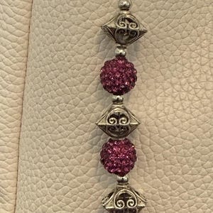 May include: Silver-toned keychain with a decorative charm. The charm features alternating sections of ornate silver filigree and sparkling, round, magenta-colored beads. The keychain is attached to a light-colored leather surface.