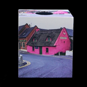 May include: A pink cottage with a thatched roof and green door sits on a street corner. The cottage has two windows and a sign that reads "The Fisherman's Cottage".