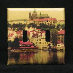 May include: A light switch cover featuring a scenic image of Prague, Czech Republic. The image shows the iconic Prague Castle, a river, and buildings with red tile roofs.
