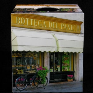 May include: A tissue box cover with a vintage Italian bakery storefront design. The storefront has a yellow awning with the text "Bottega del Pane" in gold lettering. A bicycle is parked in front of the shop.