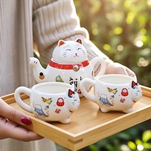 May include: A white ceramic tea set featuring a cat-shaped teapot and two matching cat-shaped teacups. The set is decorated with Japanese-style illustrations and characters, and is presented on a wooden tray.