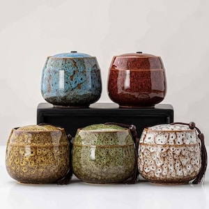 以下が含まれることがあります： 青、赤、黄、緑、白の様々な色の蓋付きセラミックジャー5個。各ジャーはユニークな斑点のある釉薬と小さなタッセルが付いています。ジャーは黒い表面に配置されています。