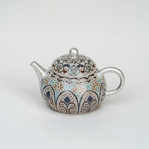Puede incluir: Una tetera de cerámica decorativa con asa, boquilla y tapa plateadas. La tetera presenta un intrincado patrón de diseños florales azules, marrones y crema. La tapa tiene un pequeño pomo plateado.