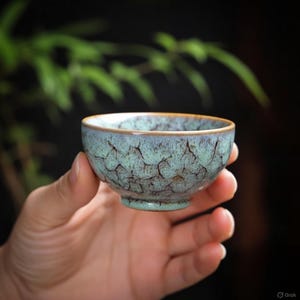 May include: A small, teal ceramic teacup with a textured surface and a brown rim. The cup is held in a hand, highlighting its design. The background is blurred, with hints of green foliage. The cup is likely for tea.