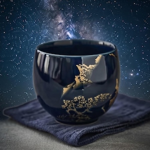 May include: A dark blue ceramic teacup with a gold landscape design. The cup sits on a dark blue cloth. The background is a starry night sky.