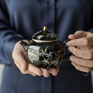 Puede incluir: Una taza de cerámica blanca con un asa negra y el texto "But First, Coffee" en negrita negra. La taza se encuentra sobre un fondo borroso, sugiriendo un ambiente acogedor.