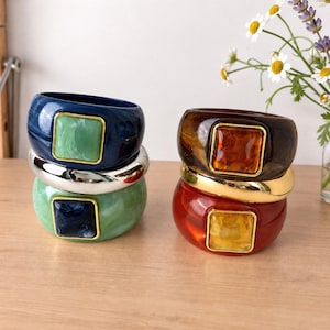 Pode incluir: Duas pilhas de três pulseiras cada. A pilha esquerda apresenta uma pulseira azul marmorizada com um quadrado verde, uma pulseira prateada e uma pulseira verde marmorizada com um quadrado azul. A pilha direita tem uma pulseira castanha marmorizada com um quadrado âmbar, uma pulseira dourada e uma pulseira vermelha marmorizada com um quadrado amarelo.