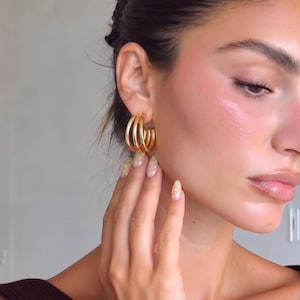 May include: A gold-toned hoop earring featuring a triple-layered design with three distinct, parallel circular bands. The earring has a polished, reflective metallic finish and a secure closure mechanism, creating a stacked appearance on the earlobe.