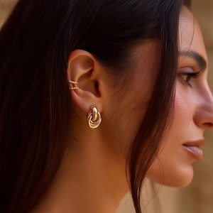 May include: Gold-coloured earrings featuring a twisted knot design. The earrings are worn on the ear of a person with dark hair. A gold ear cuff with small, sparkling stones is also visible. The background is a blurred, warm tone.
