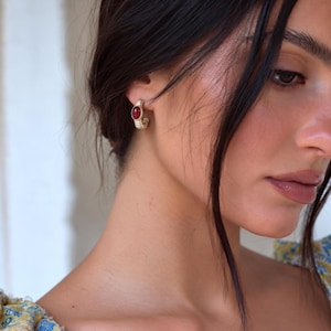 May include: A small gold-toned hoop earring featuring a textured, ribbed metal surface and a single, smooth, oval-shaped deep red gemstone set prominently at the center of the hoop, worn on the earlobe.